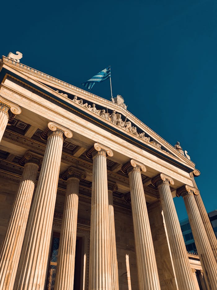 Sculpted columns of the Academy of Athens with the Greek flag under a clear sky at sunset.
